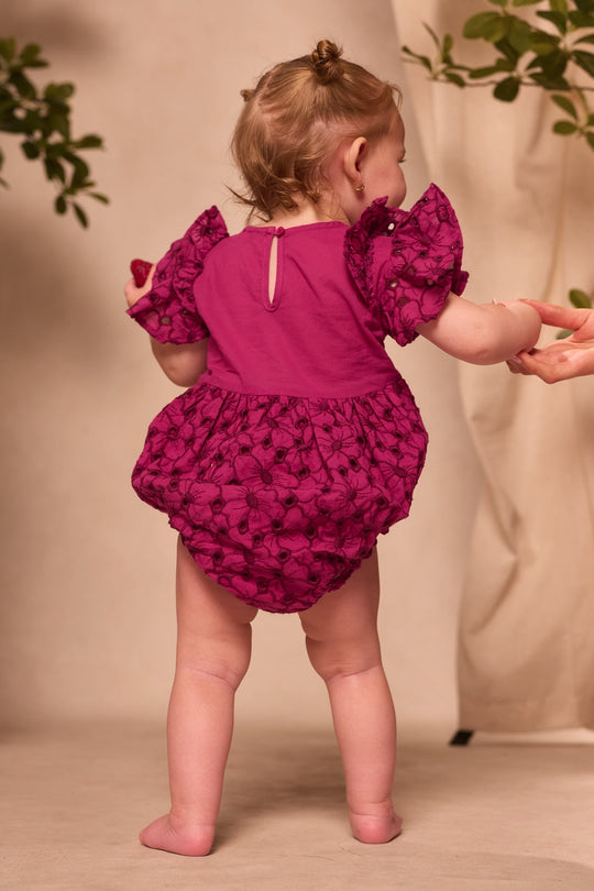 A baby stands barefoot, holding an adult’s hand and facing away from the camera, dressed in the Ivy City Co Baby Hattie Romper in Magenta—a charming embroidered cotton romper with puff sleeves and floral patterns, perfect for an adorable Easter outfit moment.