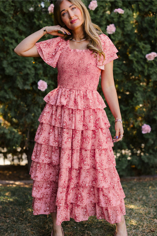 A woman with light brown hair stands outdoors, touching her head with one hand, while wearing the Grace Dress in Desert Heritage Rose by Ivy City Co—a stylish, maternity-friendly dress featuring pink floral print and tiered ruffles. Lush green bushes and pink flowers fill the background.