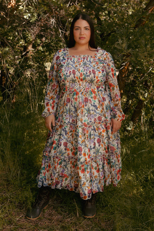 A woman stands on grass among leafy trees, wearing Ivy City Cos Selena Dress in Vivid Meadow—a floral midi dress with long sleeves, smocked bodice, and tiered ruffle skirt—paired with dark lace-up boots. She faces the camera neutrally.