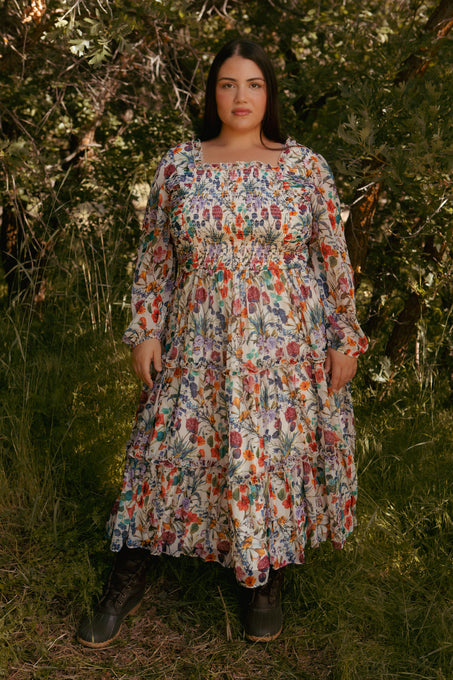 A woman stands on grass among leafy trees, wearing Ivy City Cos Selena Dress in Vivid Meadow—a floral midi dress with long sleeves, smocked bodice, and tiered ruffle skirt—paired with dark lace-up boots. She faces the camera neutrally.