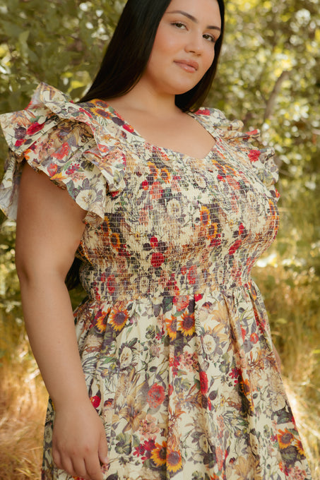 A woman wears the Hattie Dress in Rose Vine by Ivy City Co, featuring a smocked bodice and ruffled sleeves, standing outdoors among green trees and sunlight. This style is also maternity friendly.