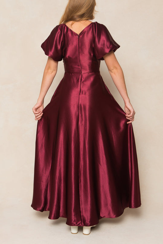 A woman is seen from the back wearing the Tessie Dress in Wine Satin by Ivy City Co, a shiny satin gown with short sleeves and a floor-length, flared hem. She holds the skirt out slightly, revealing its fitted waist—making it an ideal choice for fall bridesmaids. She completes the look with white shoes.