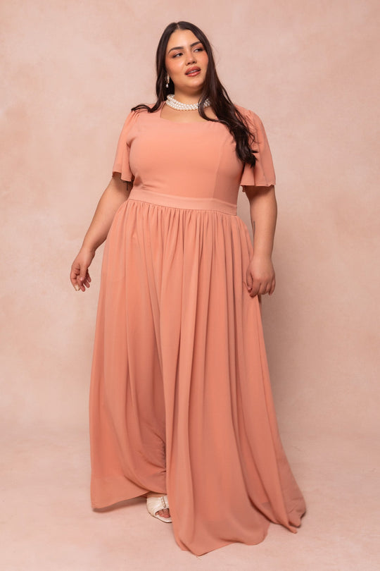 A woman stands against a soft beige background, wearing Ivy City Co’s Abbie Dress in Apricot Crush. Her long dark hair frames her face as she gazes gently to the side, accessorized with a pearl necklace and white shoes.
