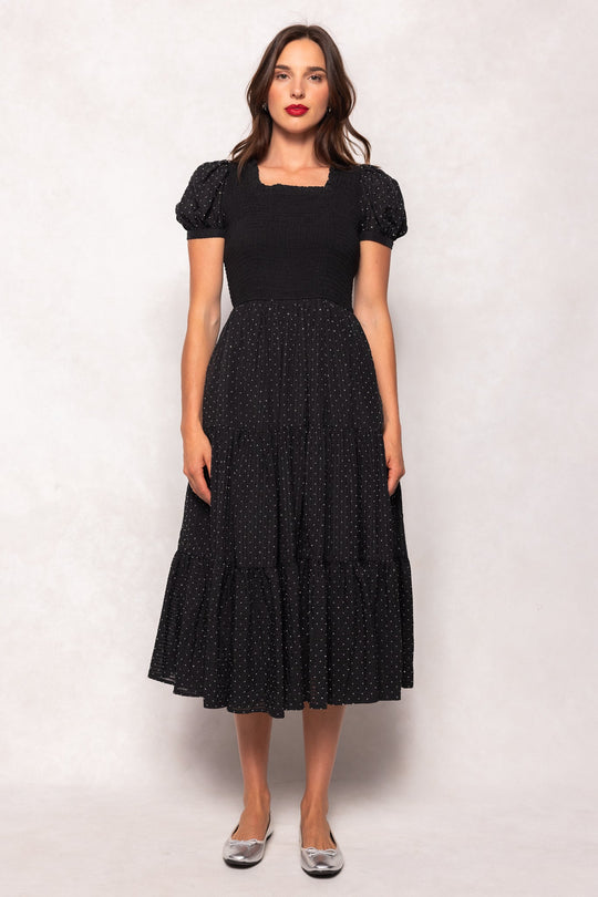 A woman stands against a pale backdrop wearing the Ivy City Co Darby Dress in Black Diamonte—a black, short-sleeve, tiered midi dress with small white polka dots—paired with silver ballet flats. Her loose, wavy brown hair and natural makeup complete the look. The Darby Dress is both stylish and maternity friendly, providing comfort without sacrificing elegance.