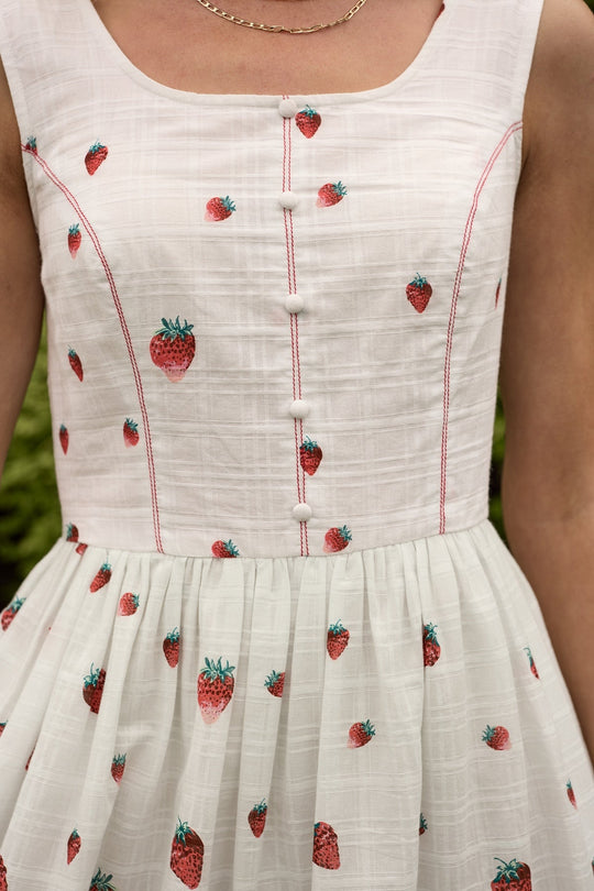 A close-up of the Sadie Dress in Strawberry Vintage Tier by Ivy City Co showcases white cotton fabric adorned with red strawberry prints, delicate pink stitching, fabric buttons down the front, and a gathered skirt. A gold chain necklace is visible around the neck, completing the charming look of this summer dress.