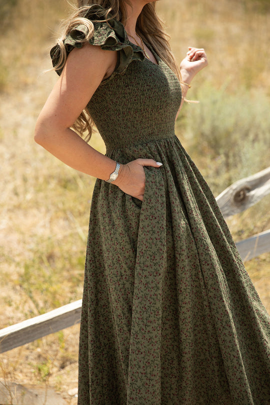 A woman in the Ivy City Co Hattie Dress in Woodland Floral, featuring a smocked bodice and ruffled sleeves, stands outdoors near a wooden fence with her hand in her pocket, surrounded by dry grass and shrubs. This effortlessly chic look is also maternity friendly.