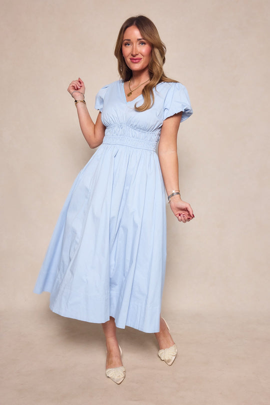 Wearing the Zella Dress in Sky Blue by Ivy City Co—a short-sleeve midi dress featuring a gathered waist—a woman stands on a beige background, smiling and posing with one hand raised. She completes her look with pointed beige shoes and gold jewelry.