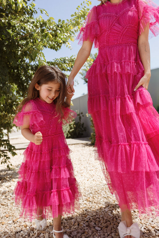 A young girl and an adult hold hands outdoors, both dressed in matching Ivy City Co Mini Whimsical Dresses in Fuchsia featuring flutter sleeves and layered tulle skirts. The girl smiles as she looks down, with sunlight streaming through the leafy green trees behind them.
