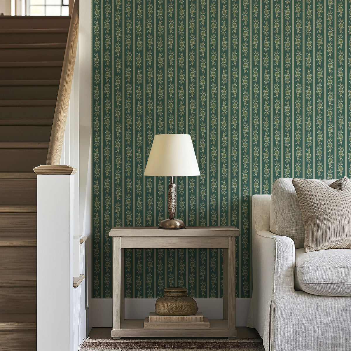 A small table with a lamp sits next to a light-colored sofa by a staircase. The wall behind is adorned with Felicity Floral Wallpaper by Loomwell Home Goods, featuring elegant white designs that add a touch of charm to the space.