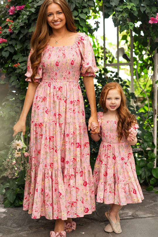 A woman and a young girl, likely mother and daughter, stand together wearing matching Ivy City Co Madeline Maxi Dresses in Pink Vintage Poppies. Holding hands and smiling amidst green plants and flowers, the woman’s nursing-friendly dress offers both comfort and effortless style—perfect for special moments together.