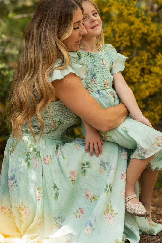 A woman and a young girl, both wearing matching cotton-linen dresses—including the charming Mini Hattie Dress in Roselyn Blue from Ivy City Co—sit together outdoors amidst yellow flowers and lush greenery, smiling and enjoying each other’s company.