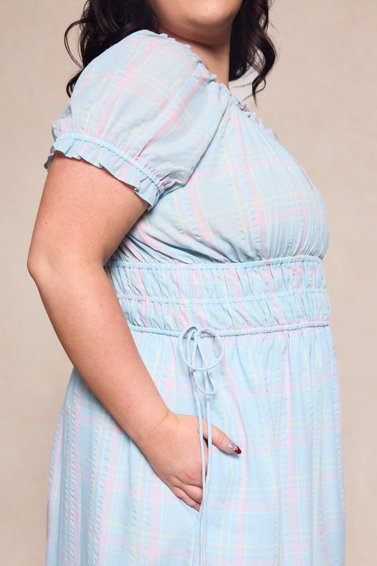 A person wearing the Ivy City Co Thalia Dress in Pastel Plaid, a light blue, short-sleeve spring dress featuring an elastic waist and side pocket, stands against a beige background. This stylish dress is designed to be maternity friendly.