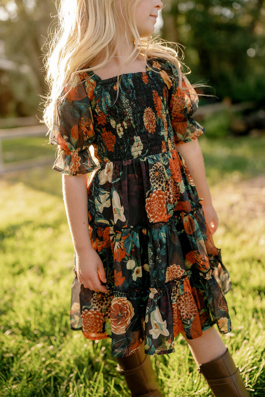 A young girl with long blonde hair wearing the Ivy City Co Mini Madeline Dress in Imogen Moody Floral and brown boots stands on grass in sunlight, trees softly blurred in the background.