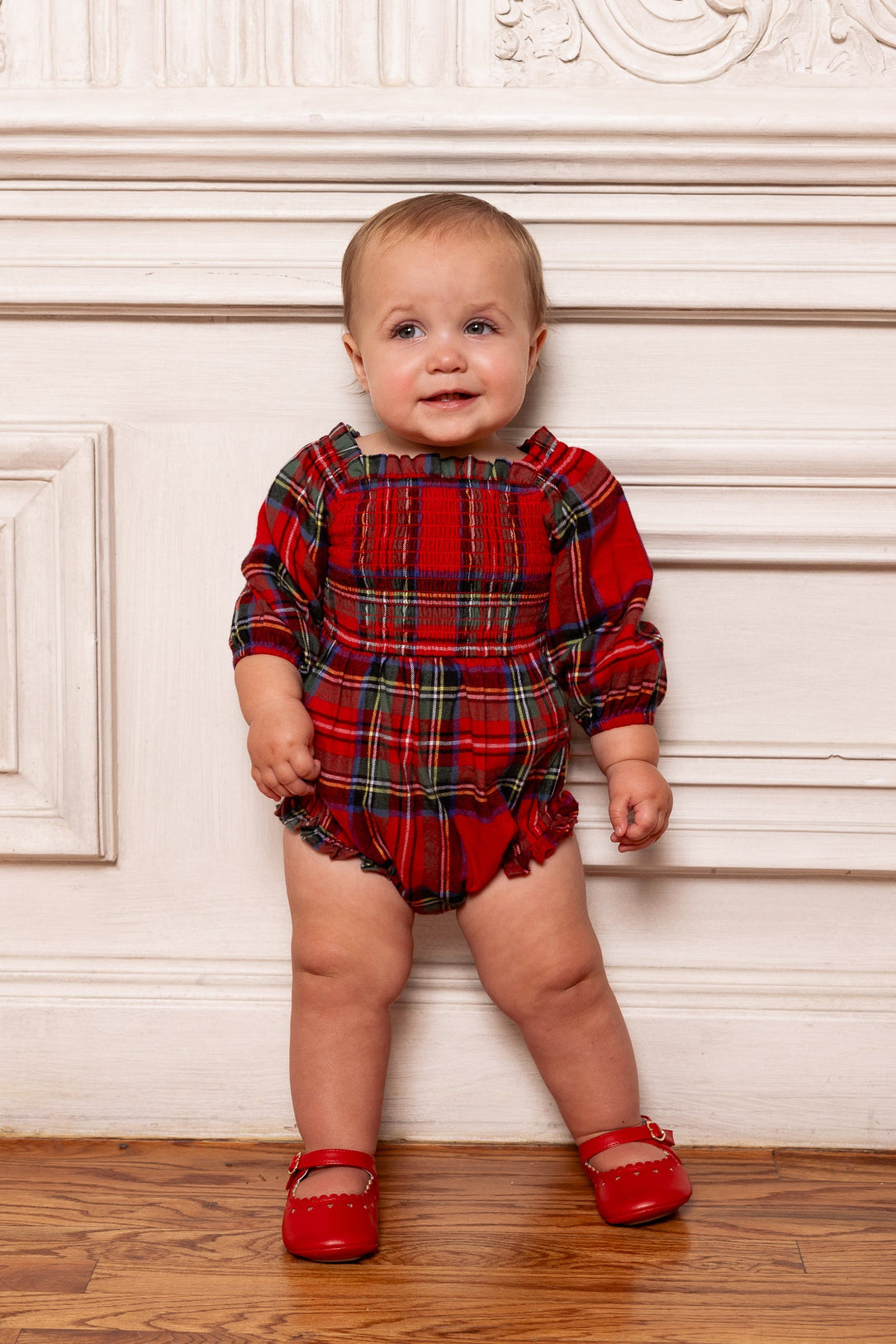 A baby wearing the Ivy City Co Baby Madeline Romper in Holiday Plaid and red shoes stands against a white paneled wall on a wooden floor, looking upward and smiling—making for the perfect baby holiday outfit.