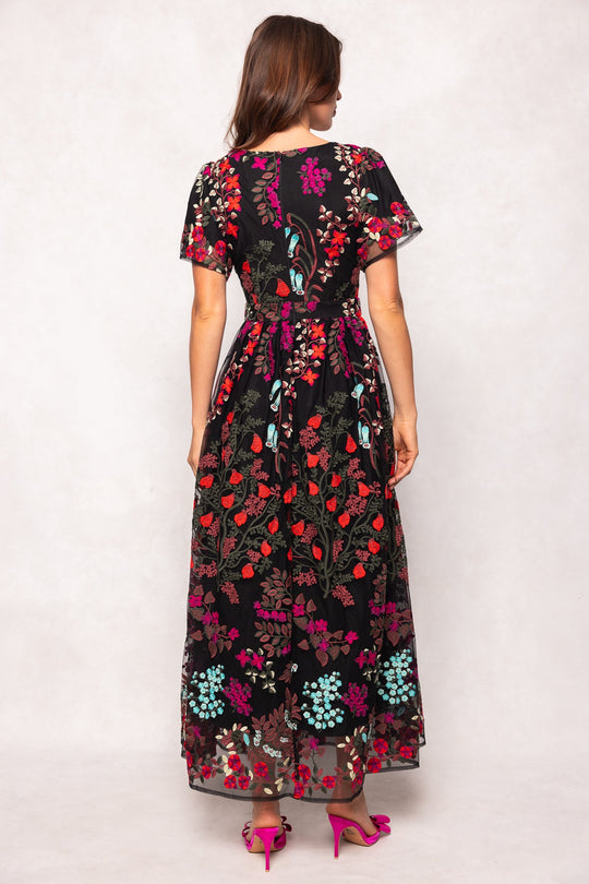 A woman stands facing away, wearing the Flora Dress in Moonlit Blossom by Ivy City Co—a long black dress featuring vibrant embroidered floral patterns. The dress is styled with short sleeves and a fitted waist. She has brown hair and pairs the look with bright pink heeled sandals.