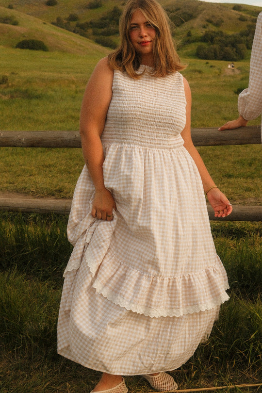 Wearing the Presley Sleeveless Dress in Cream Gingham by Ivy City Co, a woman stands outdoors beside a wooden fence with rolling green hills behind her. The maxi length and smocked bodice of her dress enhance the breezy charm of the scene.
