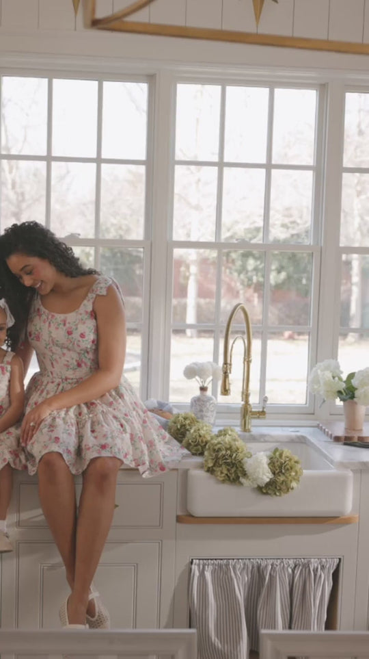 A woman wearing the Helene Dress in Vibrant Chintz by Ivy City Co sits on a kitchen counter beside a sink adorned with flowers. Large windows behind her fill the space with natural light, and a vase of white blooms rests on the countertop.