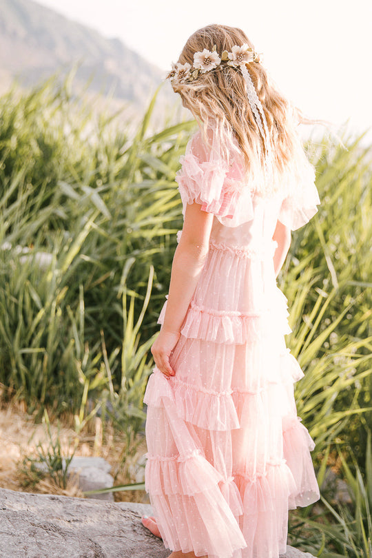 A young girl with wavy blonde hair wears the Ivy City Co Mini Whimsical Maxi Dress in Blush and a flower crown, standing barefoot on a rock with tall green grass in the background.