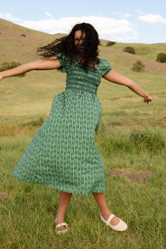 Wearing the Ivy City Co Sunday Best Dress in Pine Felicity Floral—designed to be nursing and maternity friendly—a person twirls joyfully in a grassy field, with rolling hills and a partly cloudy sky behind them.