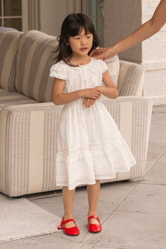 A young girl wearing the Ivy City Co Mini Palermo Dress in White and red shoes stands indoors near a striped sofa, as an adult hand reaches toward her face.