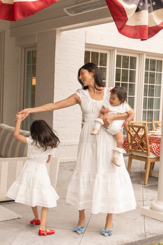 A woman wearing the Palermo Dress in White by Ivy City Co dances with a young girl in a matching outfit, gently holding a baby. Their maternity-friendly Palermo Dresses create a sweet scene on the patio, adorned with patriotic bunting and red-checkered tablecloths in the background.