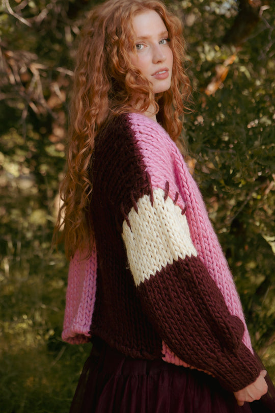 A person with long curly red hair stands outdoors amid green foliage, looking over their shoulder while wearing the Marissa Sweater in Pink & Red by Ivy City Co.
