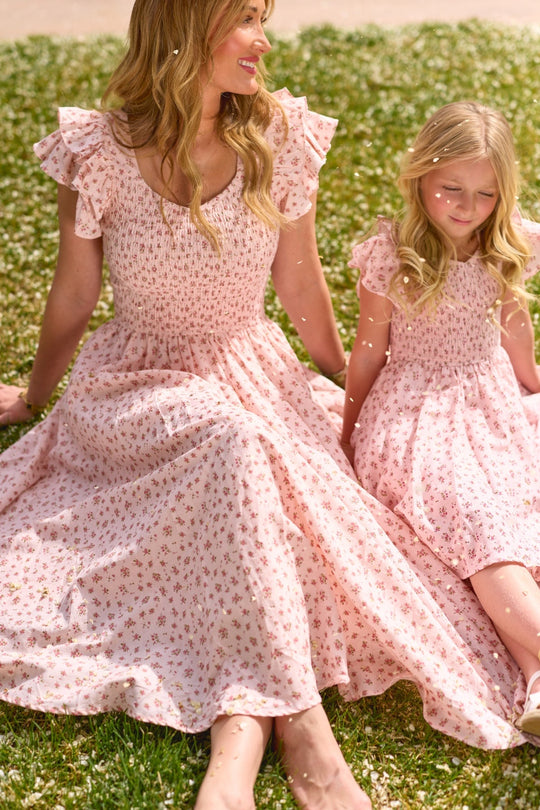 A woman and a young girl sit on the grass in matching Ivy City Co Hattie Dresses in Madeline Pink. They smile and look relaxed, enjoying a sunny day outdoors—perfectly dressed for the occasion in their coordinated floral ensembles.