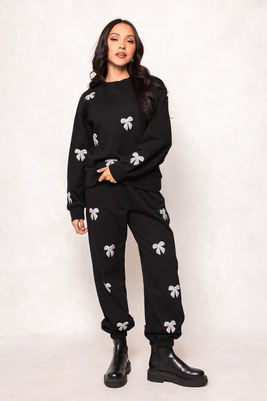 A woman in a black sweatshirt and Ivy City Co Micah Jogger Sweatpants in Black, adorned with silver and rhinestone bows, wears black boots and stands against a light neutral background.