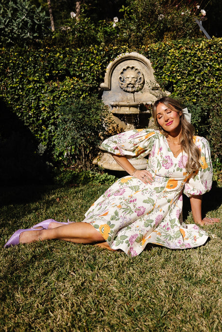 A woman in the Ivy City Co Leah Dress in Multicolor Floral and lavender heels sits on the grass, leaning on one hand, with greenery and a decorative stone fountain behind her.