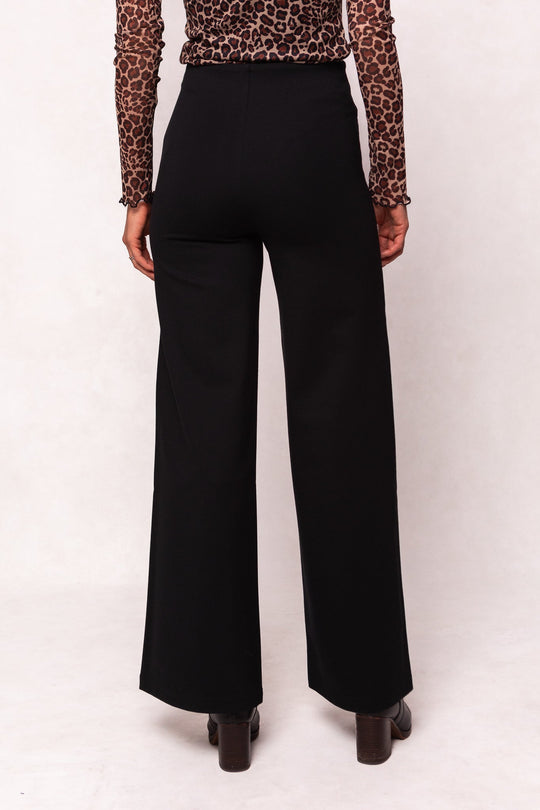 A person is standing facing away from the camera against a plain light background, wearing the Alyssa Pop Ponte Pants in Black by Ivy City Co—a high-waisted, wide-leg style that’s perfect for day-to-night wear—paired with a long-sleeve leopard print top.