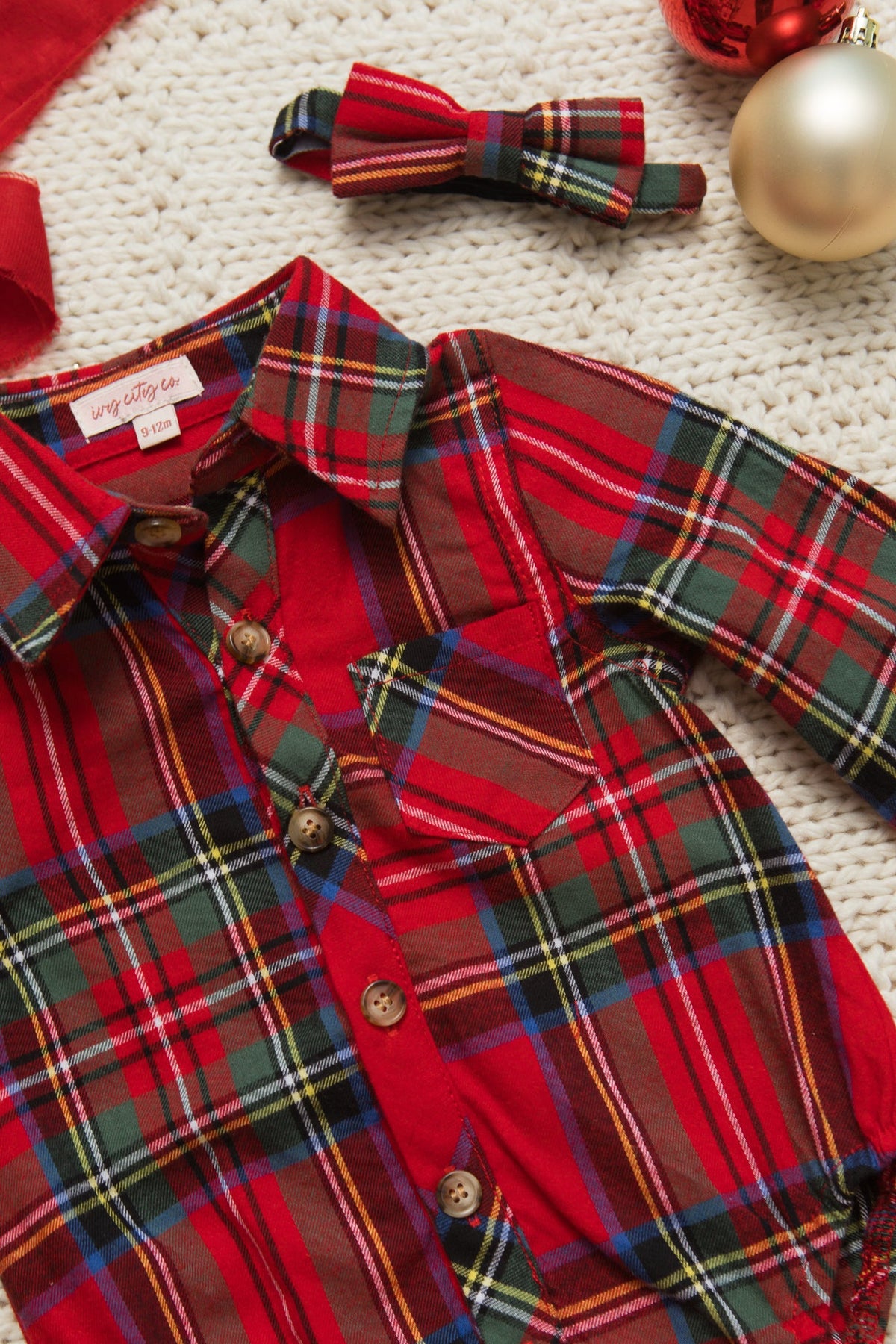 The Ivy City Co Baby Boys John Romper in Holiday Plaid, a festive red, green, and yellow button-up romper with a matching bow tie, is displayed on a white knit surface beside red and gold Christmas ornaments.
