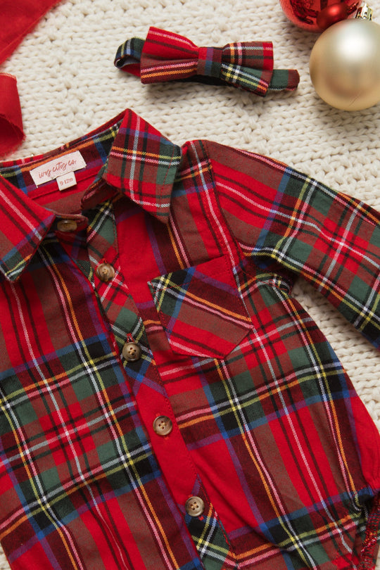 The Ivy City Co Baby Boys John Romper in Holiday Plaid, a festive red, green, and yellow button-up romper with a matching bow tie, is displayed on a white knit surface beside red and gold Christmas ornaments.