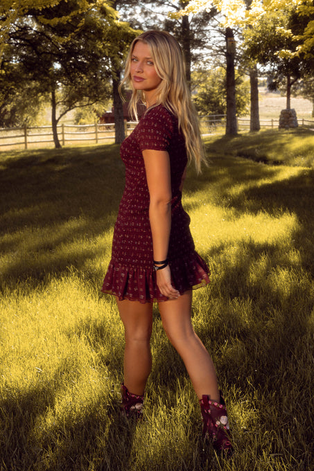 A woman with long blonde hair wears the Ivy City Co Elaine Dress in Mulberry Vine and matching boots, standing on sunlit grass with trees and a wooden fence in the background.
