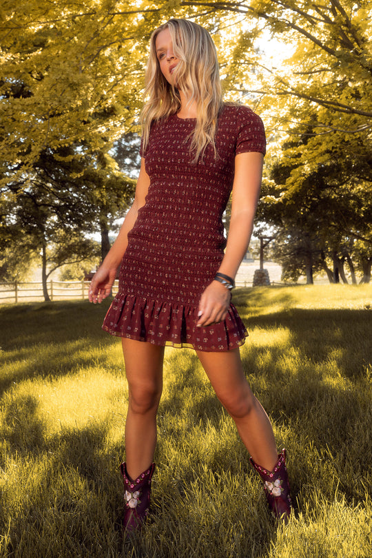 A woman stands in sunlight on grass, wearing the Ivy City Co Elaine Dress in Mulberry Vine—a short maroon floral piece with a ruffled hem—paired with matching maroon cowboy boots. Yellow-green trees and a wooden fence complete the rustic scene.