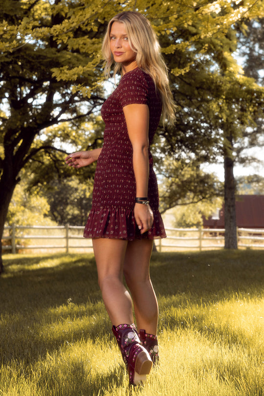 A woman with long blonde hair stands on grass outdoors, turning to look over her shoulder as sunlight filters through the trees. She wears the Ivy City Co Elaine Dress in Mulberry Vine and matching boots.
