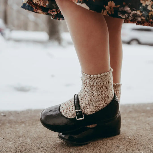 A person wearing cream knit socks and Zimmerman Shoes’ Mini Mary Janes in Black Patent stands on a snowy sidewalk, with part of a floral skirt visible.