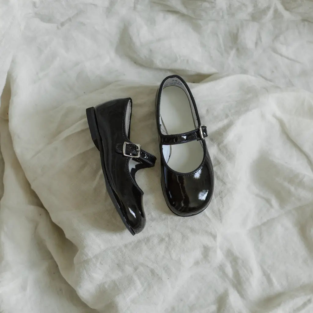 A pair of Zimmerman Shoes Mini Mary Janes in Black Patent, featuring buckled straps, displayed on a crumpled white fabric background.