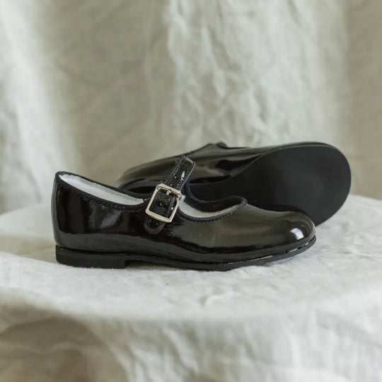 The Mini Mary Janes in Black Patent by Zimmerman Shoes are displayed on a white fabric surface against a soft, neutral background, showcasing their shiny black patent leather and classic buckle strap.