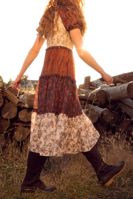 Person wearing a patterned dress standing near a stack of logs.