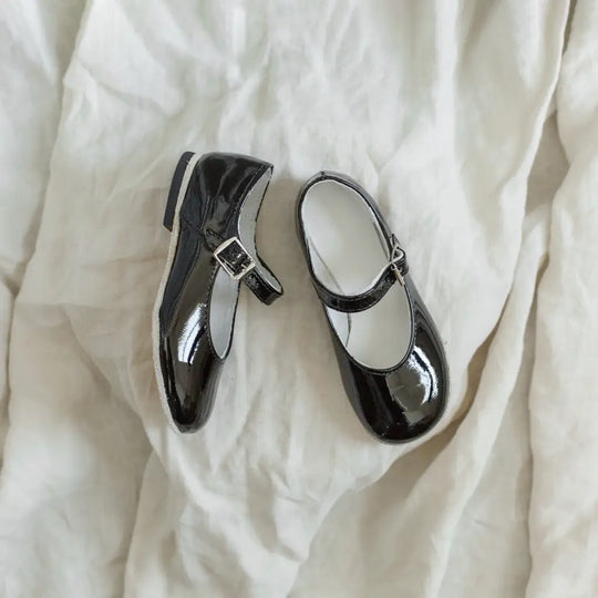 A pair of Zimmerman Shoes’ Mini Mary Janes in Black Patent, featuring adjustable ankle straps and buckles, displayed on a crumpled white fabric background.
