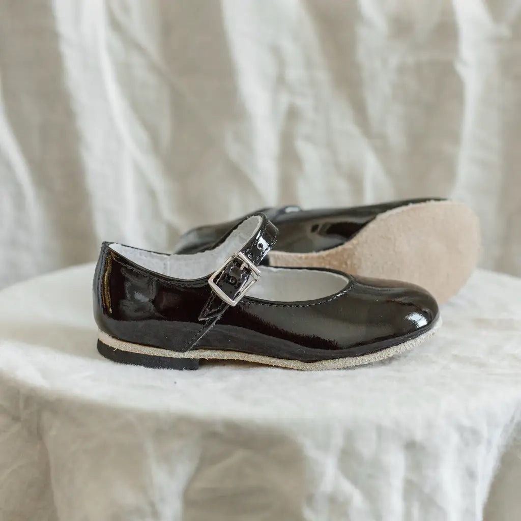 A pair of Zimmerman Shoes Mini Mary Janes in Black Patent, featuring an adjustable ankle strap with buckles, are displayed on a white textured surface against a light-colored fabric background—perfect as toddler walking shoes.