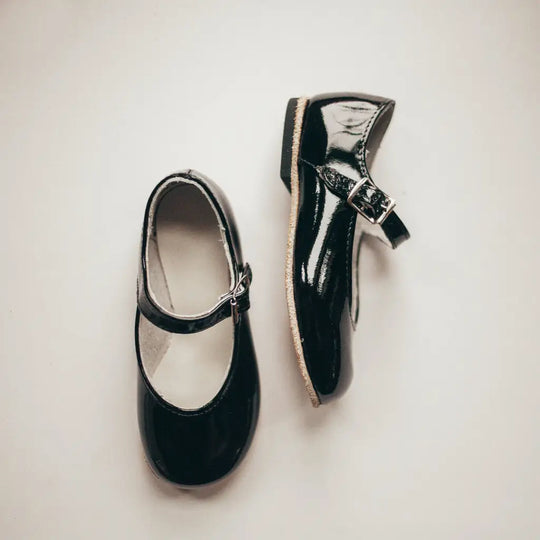 A pair of Zimmerman Shoes Mini Mary Janes in Black Patent, featuring adjustable ankle straps, is displayed on a light, neutral background.