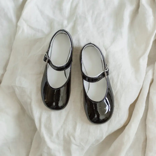 A pair of Zimmerman Shoes’ Mini Mary Janes in Black Patent, featuring buckles, are placed side by side on a soft, crumpled white fabric background.