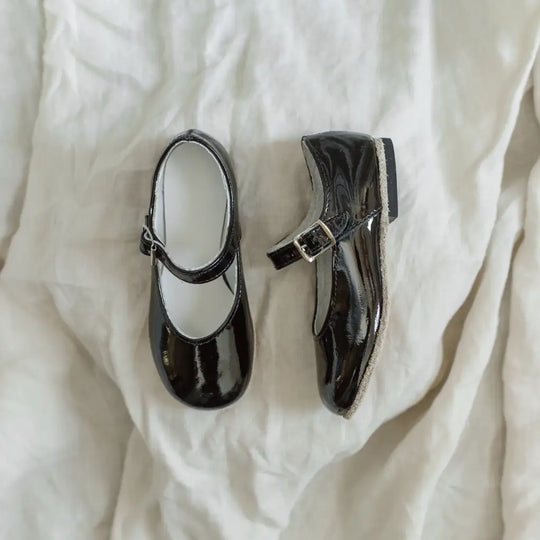 A pair of Mini Mary Janes in Black Patent by Zimmerman Shoes, featuring a single buckle strap and shiny black patent leather, displayed on a soft, crumpled white fabric background.