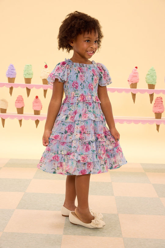 A young girl in the Ivy City Co Mini Winnie Dress in Peony Blossoms, with its smocked bodice and peony floral print, smiles while standing on a tiled floor. Behind her, colorful ice cream cone decorations adorn a pastel-colored wall.