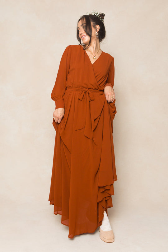 A woman wearing the Ivy City Co Andie Dress in Amber Chiffon—a long-sleeve, rust-orange wrap maxi dress with a waist tie—is holding part of the skirt while smiling and looking to the side, with flowers in her hair and standing against a beige background.