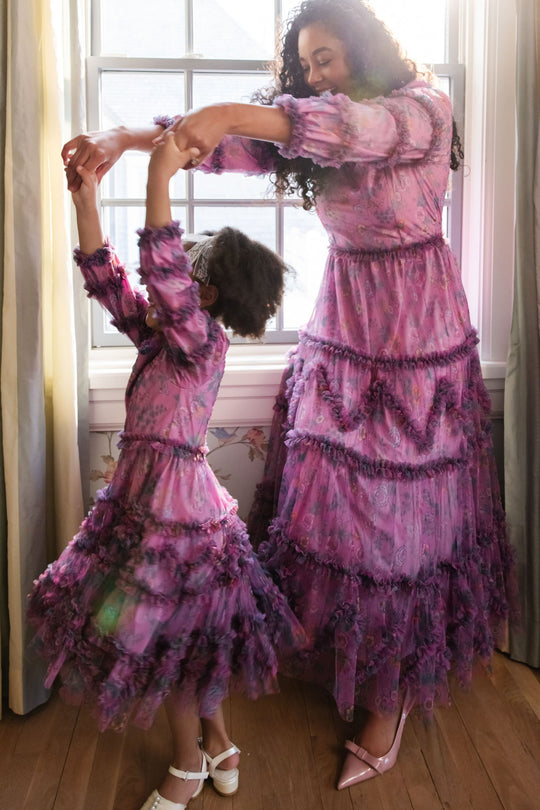 A woman and a young girl, both dressed in matching Ivy City Co Catherine Dresses in Climbing Vine Bloom, dance together in a sunlit room beside a large window adorned with lush greenery.