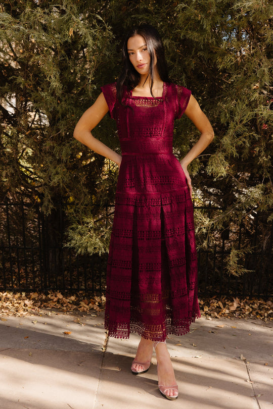 Wearing the Amory Dress in Burgundy by Ivy City Co, a woman stands on a sidewalk with her hands on her hips, posing in front of leafy trees. The lace midi dress features a square neckline and is paired perfectly with open-toe heels.