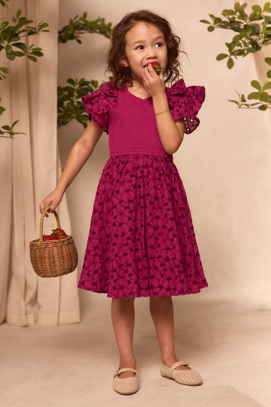 A young girl wearing the Ivy City Co Mini Hattie Dress in Magenta enjoys a strawberry while holding a basket of strawberries. She stands indoors amid green plants, smiling as she looks to the side.