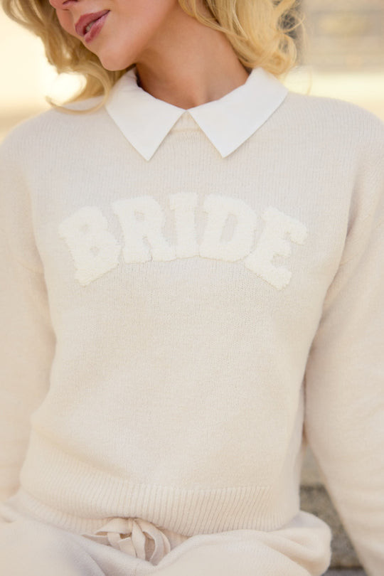 A woman wearing the Ivy City Co Bride Sweater in Oatmeal, featuring the word “BRIDE” in textured letters on the front, layered over a white collared shirt. Her face is partially visible.